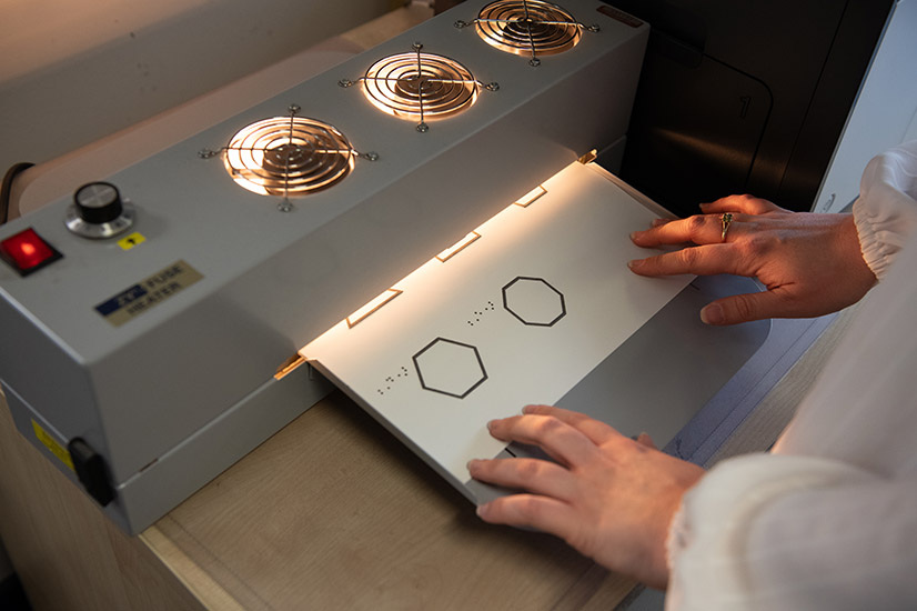 The image shows a pair of hands operating a tactile graphics embosser. The hands are gently guiding a sheet of paper or plastic into the machine. The sheet contains raised geometric shapes, such as hexagons, and Braille markings. The machine has three visible fan-like elements at the top, likely to regulate heat.