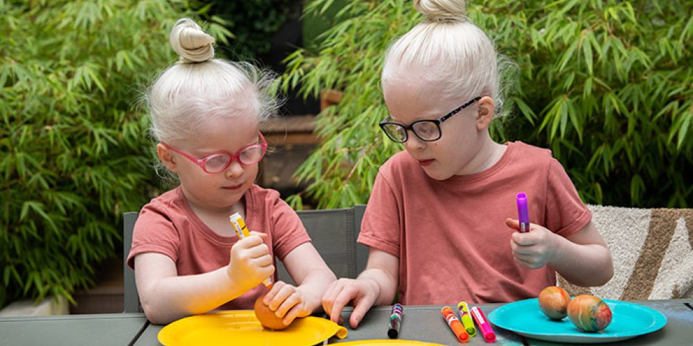 Image showing two young children who are interacting as they decorate eggs, possibly for Easter. The image shows them using coloured pen and plates in bright yellow and turquoise hues.