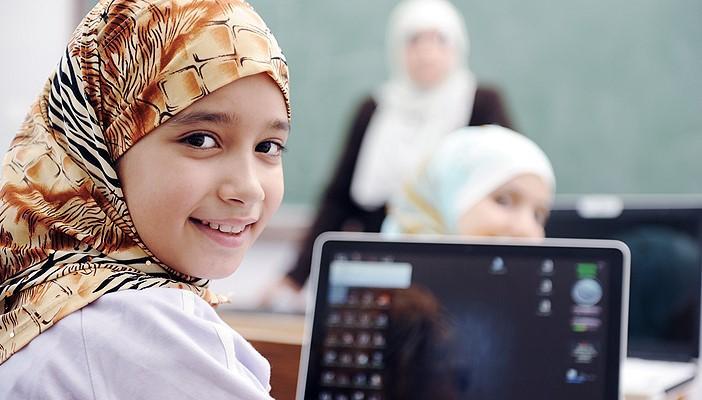 A pre-teen girl in a headscarf, smiling with a laptop on a desk, a teacher is at front of class in background.